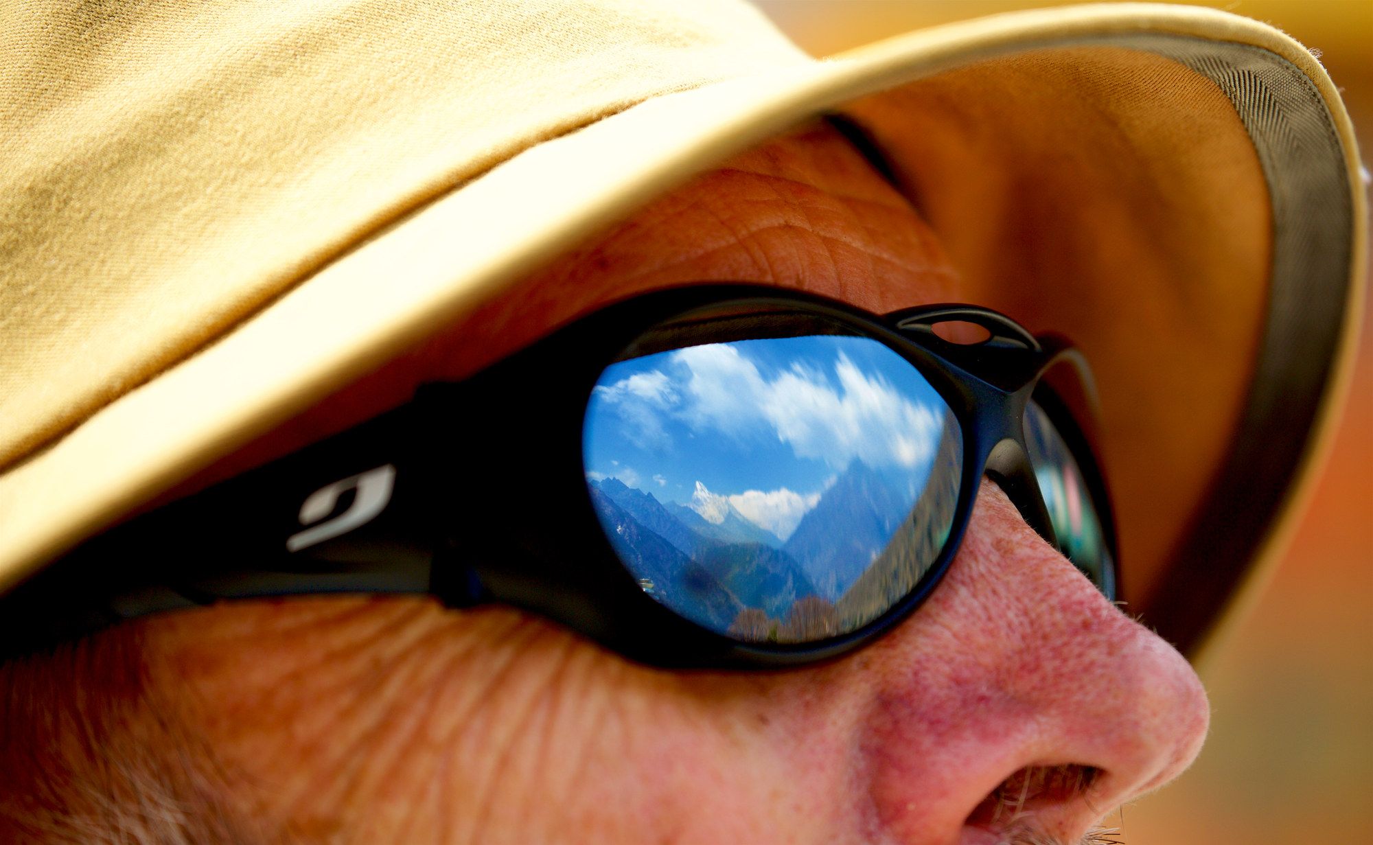 Extreme close-up of a face in a sun hat, sky and land reflected in the sunglasses