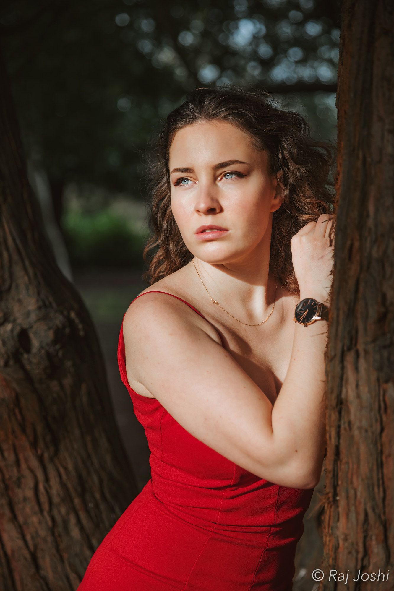 A woman in a red dress leaning against dark tree bark in soft light