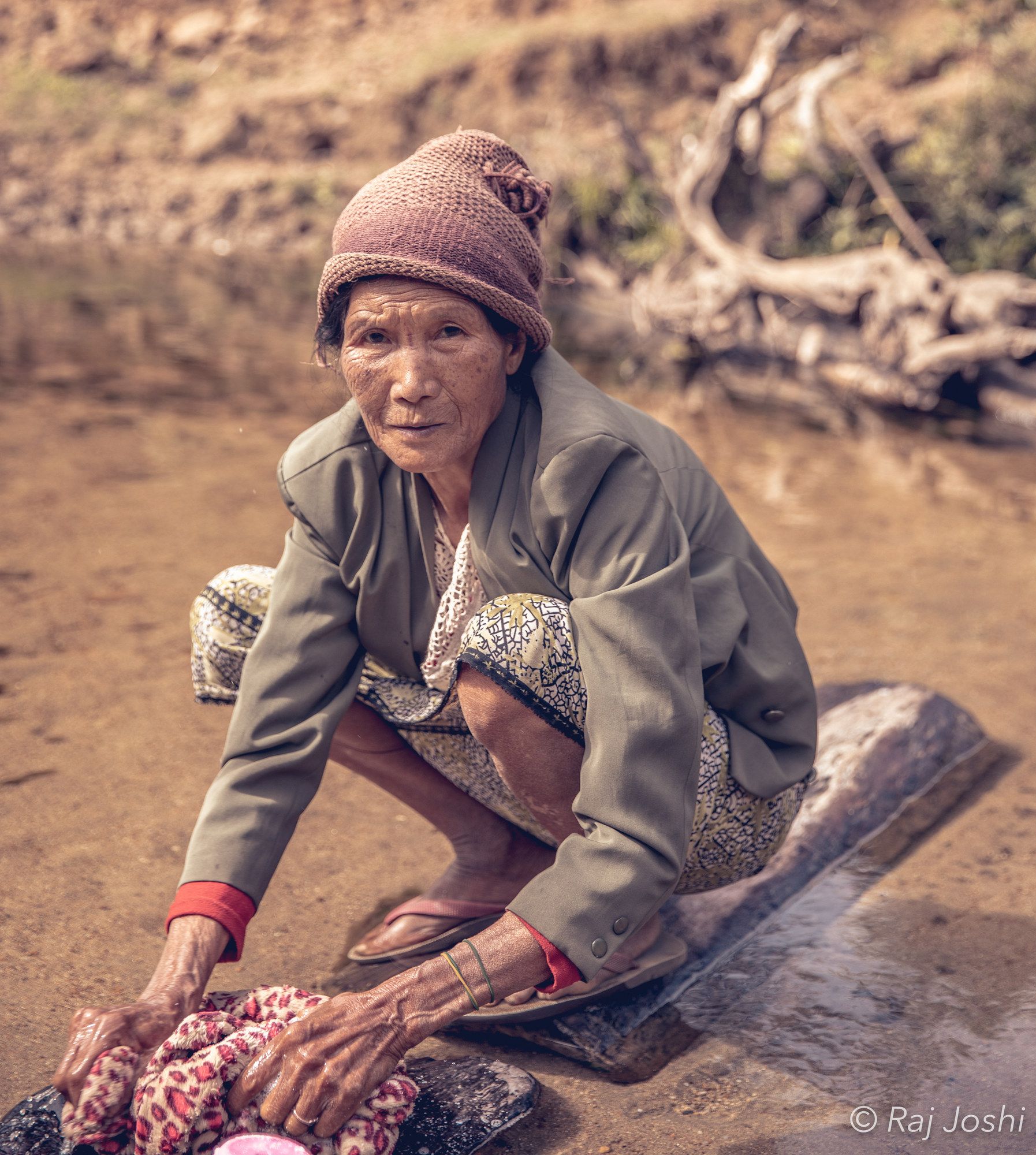 A woman in a pink hat resting on the ground beside a river camp at day’s end